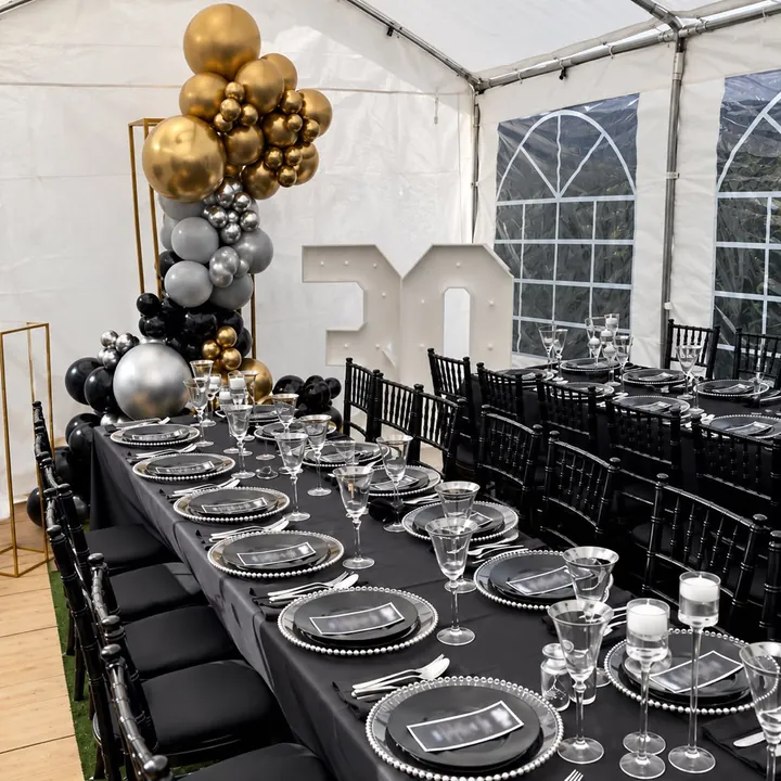 A 30th birthday party table setting with black chairs, black and silver tableware, a gold and silver balloon display and an illuminated 30