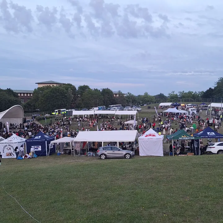 An outdoor festival field with a stage canopy, rows of trader tents and a crowd