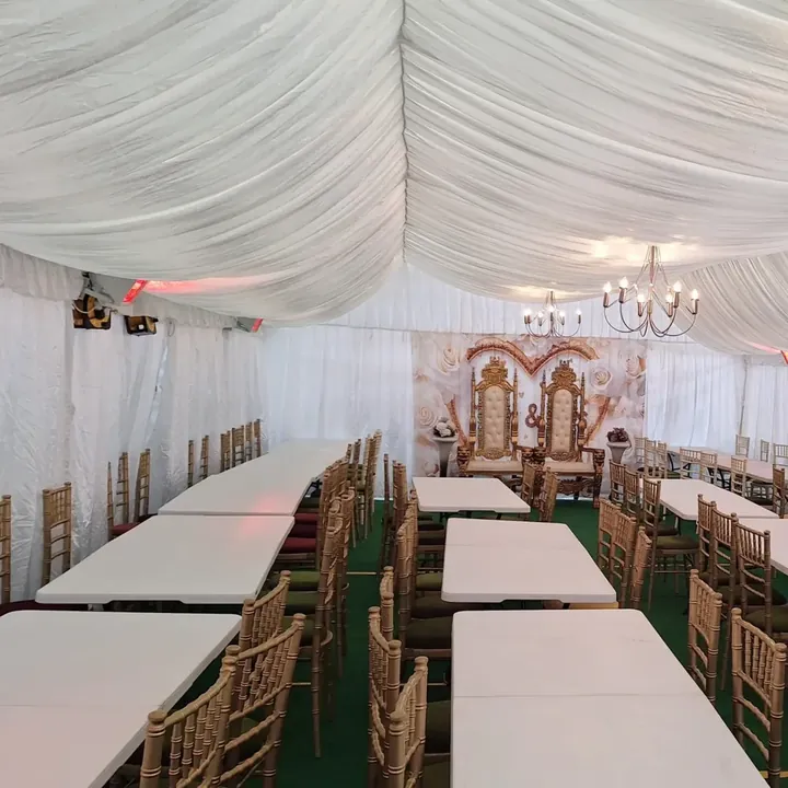 A lined marquee with banquet tables, gold chairs, chandeliers and two gold throne chairs before a floral backdrop