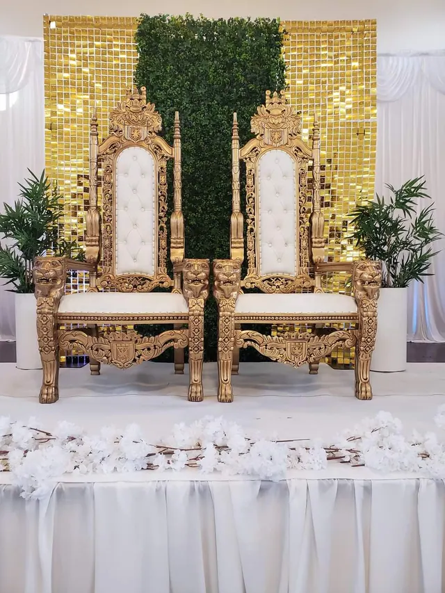 A pair of gold and white throne chairs with a decorative gold and green backdrop