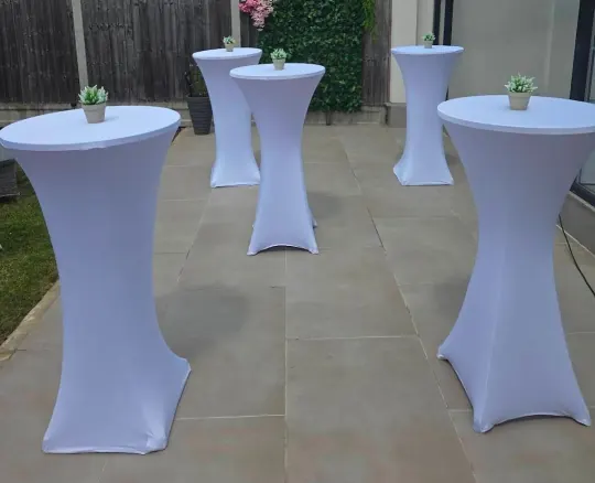 Five poseur tables dressed in white stretch covers, arranged on a paved garden patio with a potted plant on each top
